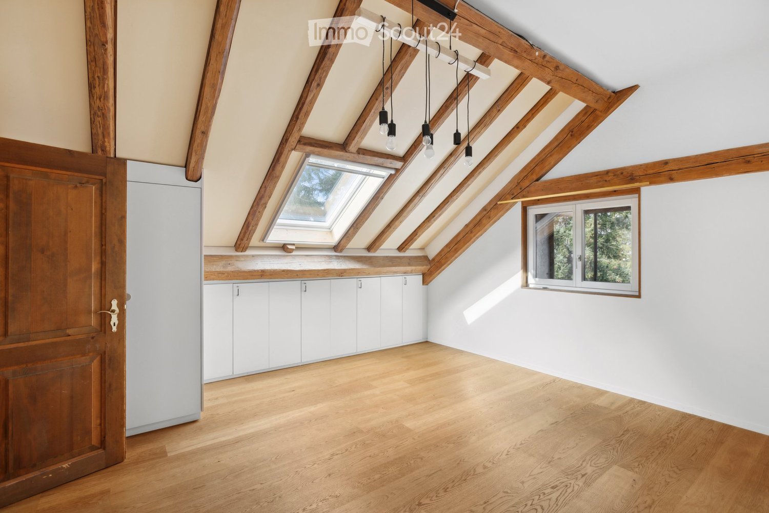 Bright, empty attic room with wooden beams, skylight, and white cabinetry. Wooden floor and white walls.