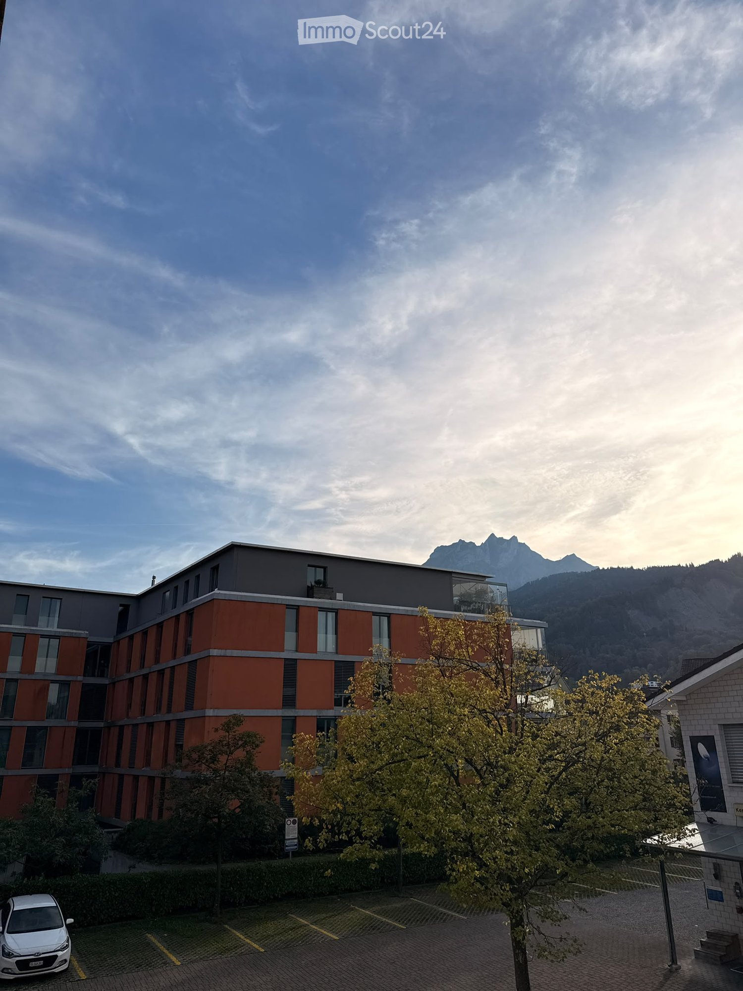 Modern multi-story building with orange and gray facade, large windows, and a parking area in front, with trees and a scenic mountain view in the background.
