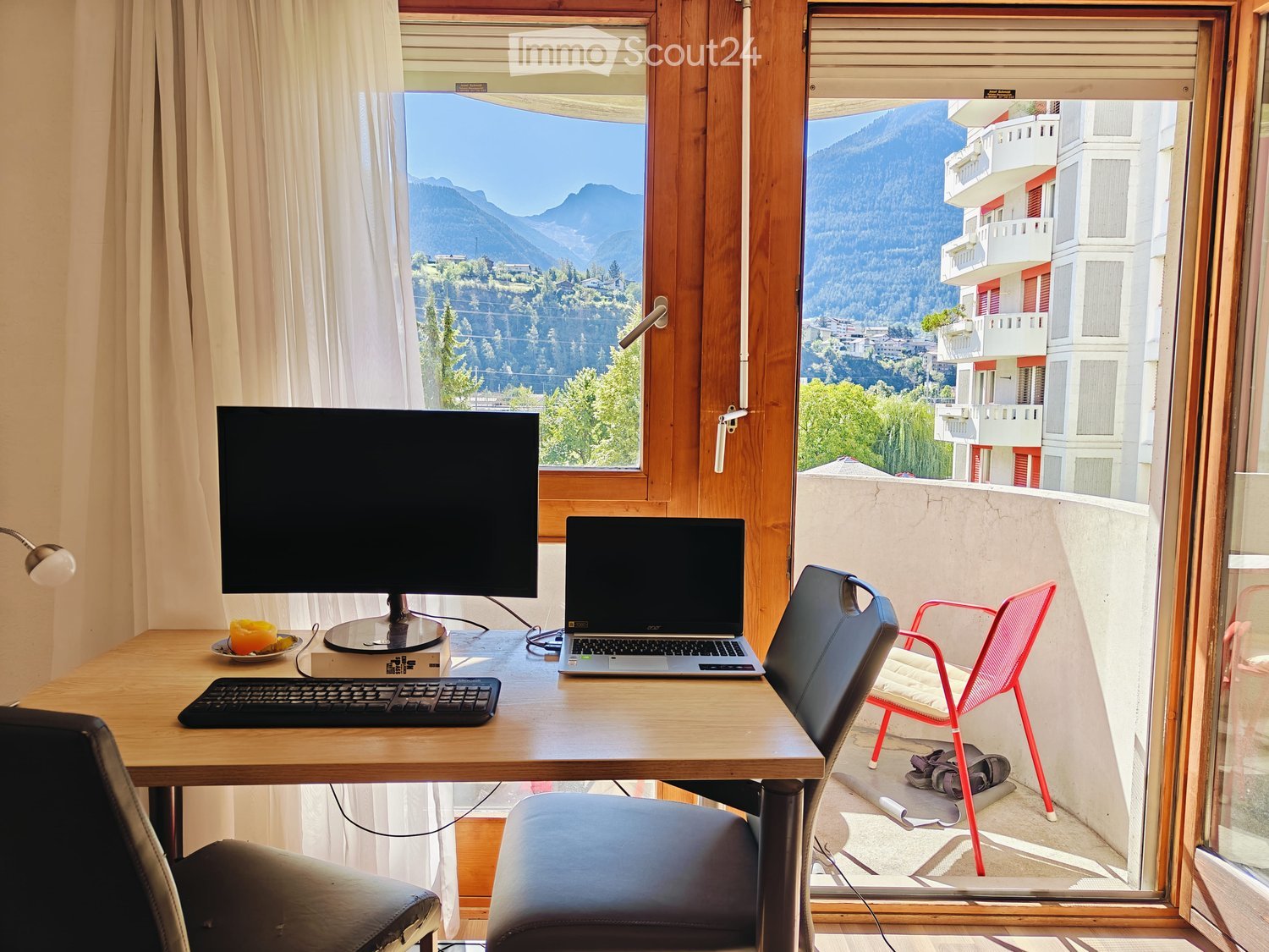 A furnished room with a desk, laptop, screen, keyboard, and chairs, overlooking a scenic mountain view.