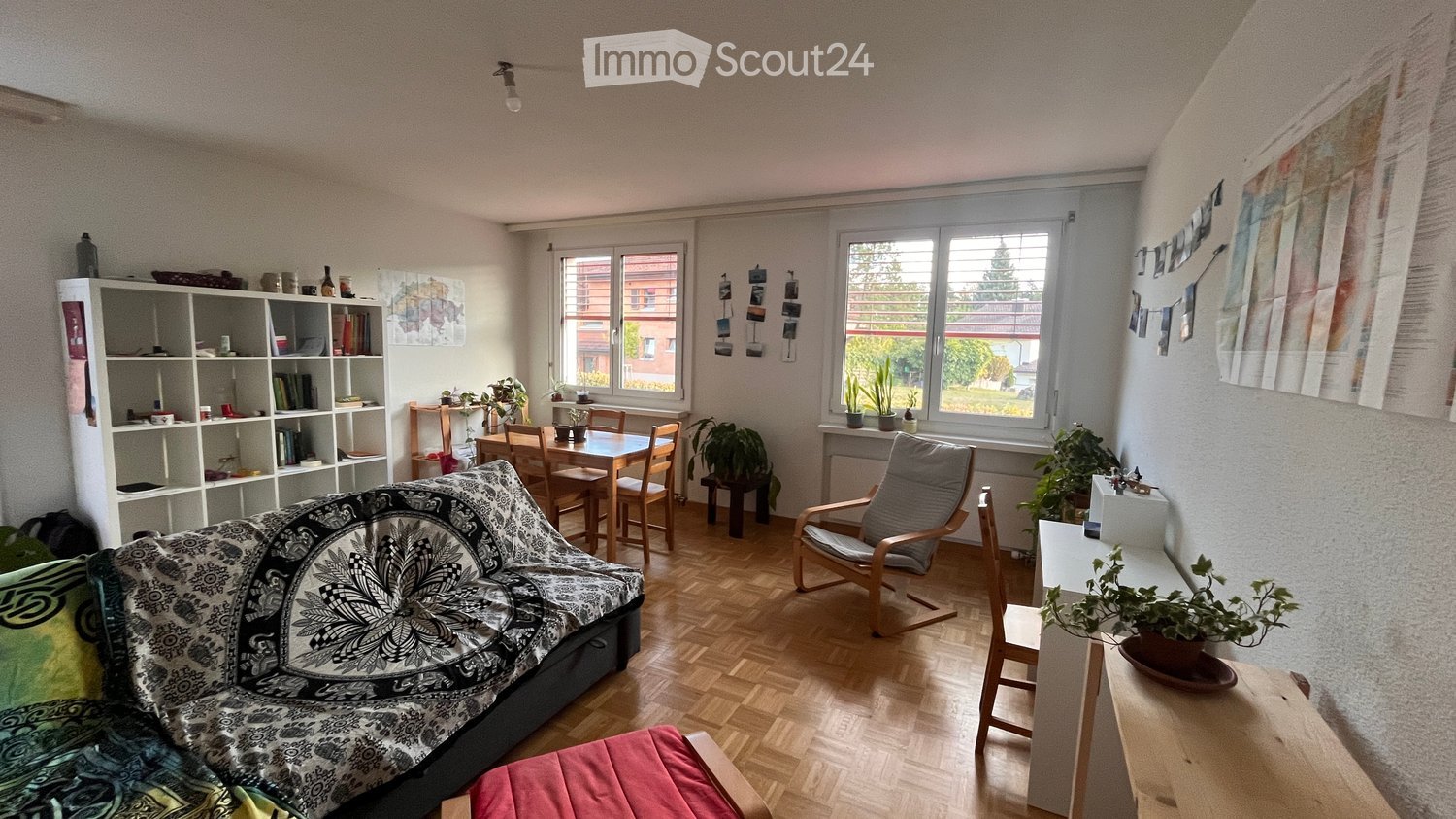 A cozy living room with a patterned sofa, wooden dining table, bookshelf, and potted plants. The room features large windows with a view of the outside and a mix of modern and rustic furniture.