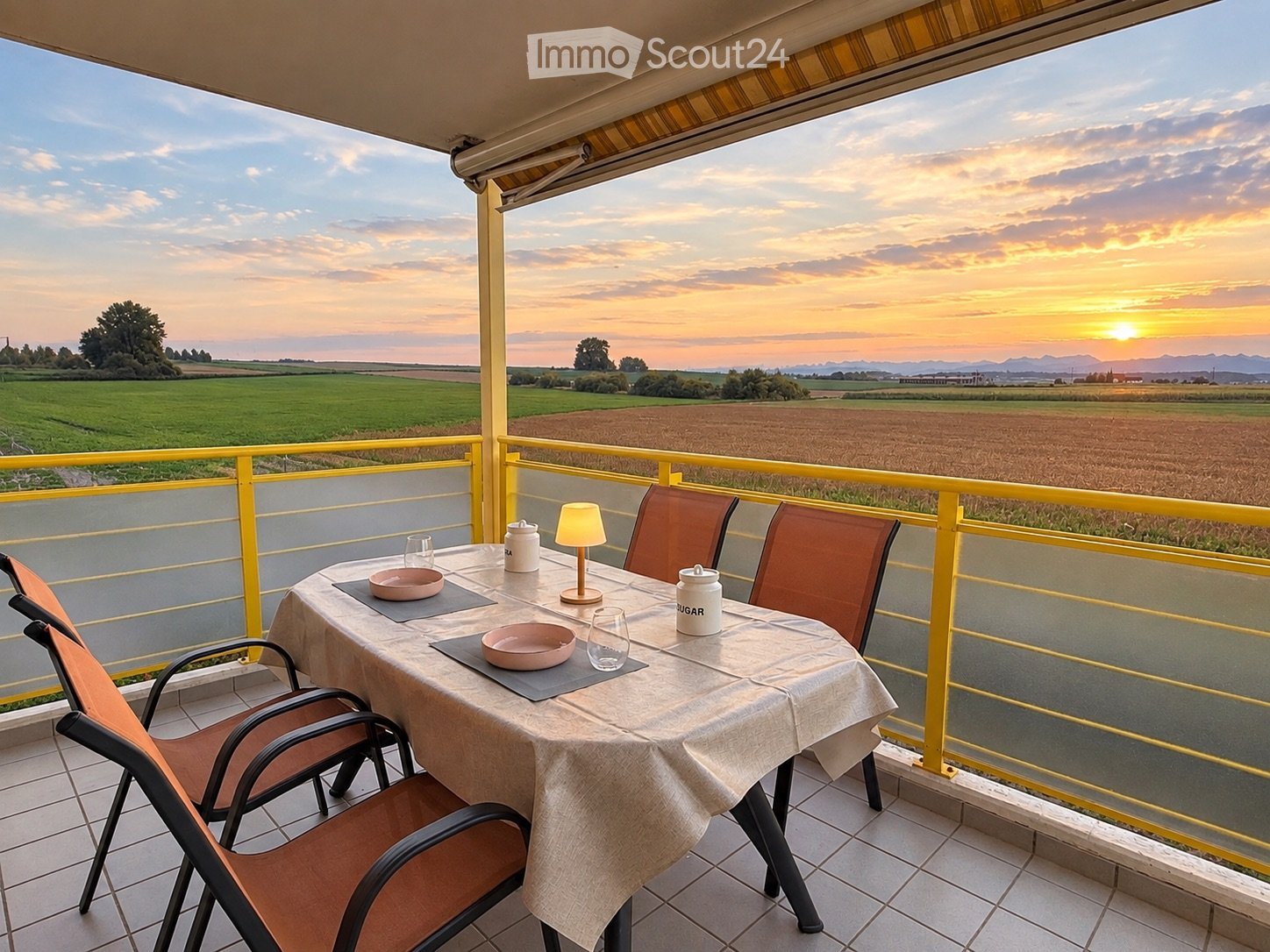 Balcony with a table set for two, featuring a lamp, cups, and bowls, overlooking a scenic countryside view at sunset.