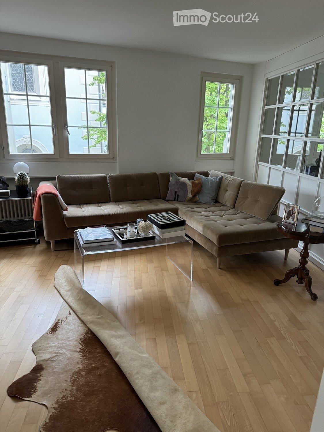 Modern living room with a large sectional sofa, glass coffee table, books, decorative cushions, a cowhide rug, and a small table with a lamp and picture frame.