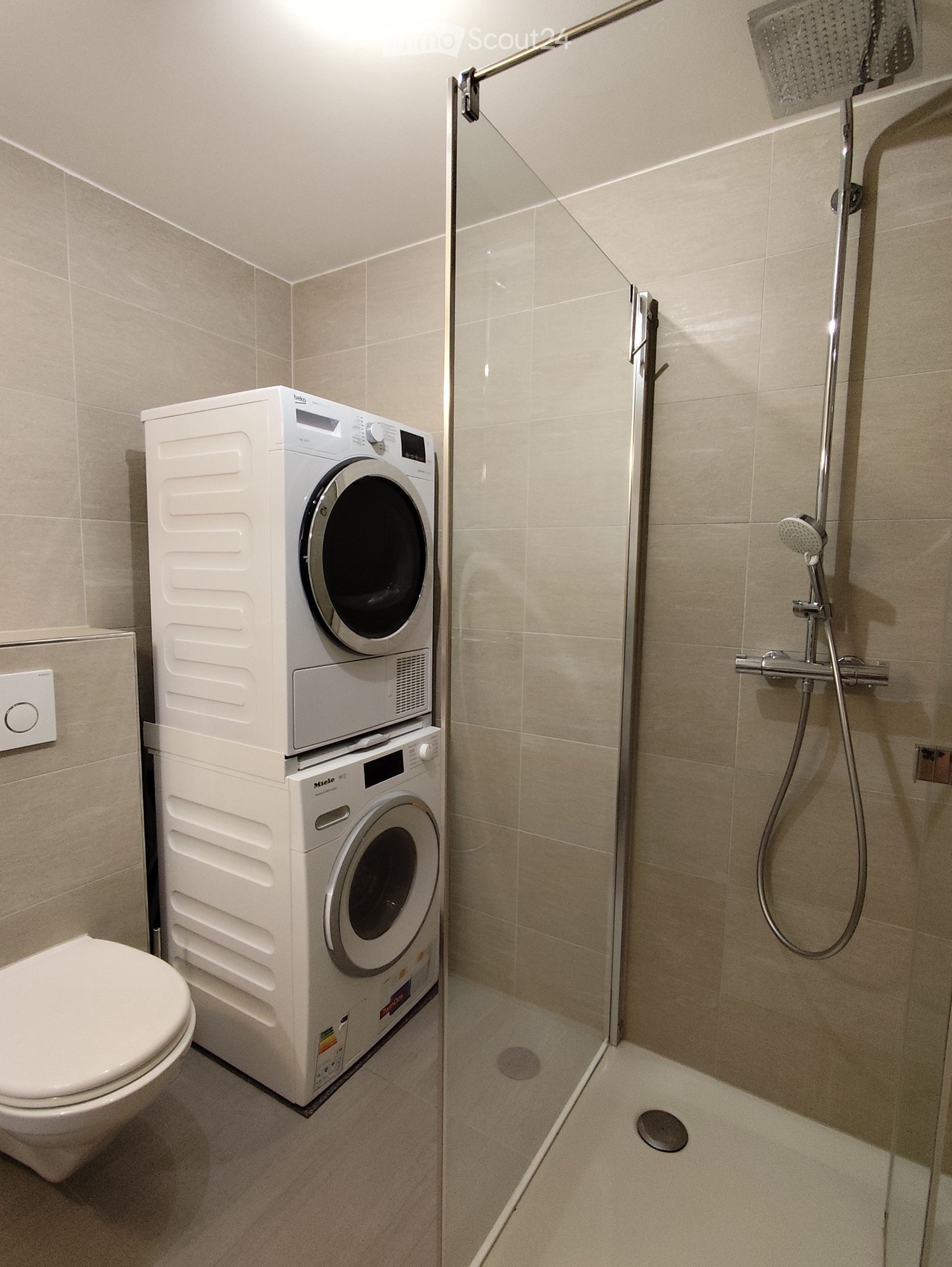 Modern bathroom with a toilet, stacked washing machine, and a glass-enclosed shower with a handheld showerhead.