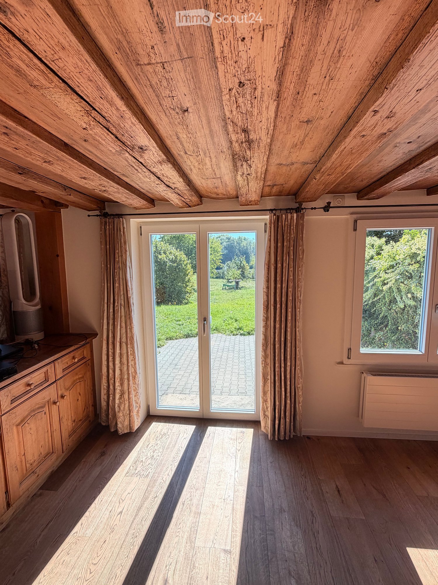 Bright attic room with wooden beams, large sliding glass doors leading to a patio, wooden flooring, and a small cabinet with a speaker on top.