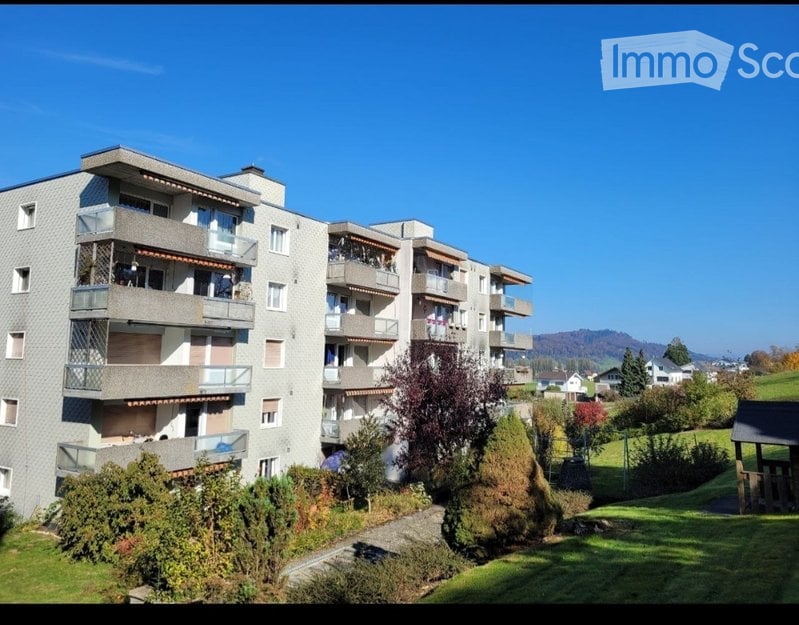 Multi-story apartment building with multiple balconies overlooking a green, hilly landscape with distant mountains.