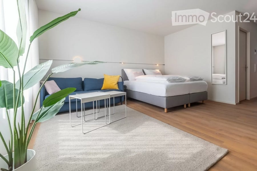 Modern, minimalist room with a blue sofa, white coffee tables, a large bed, and a leafy plant in a pot. The space features light-colored walls, wooden flooring, and a large mirror on the wall.