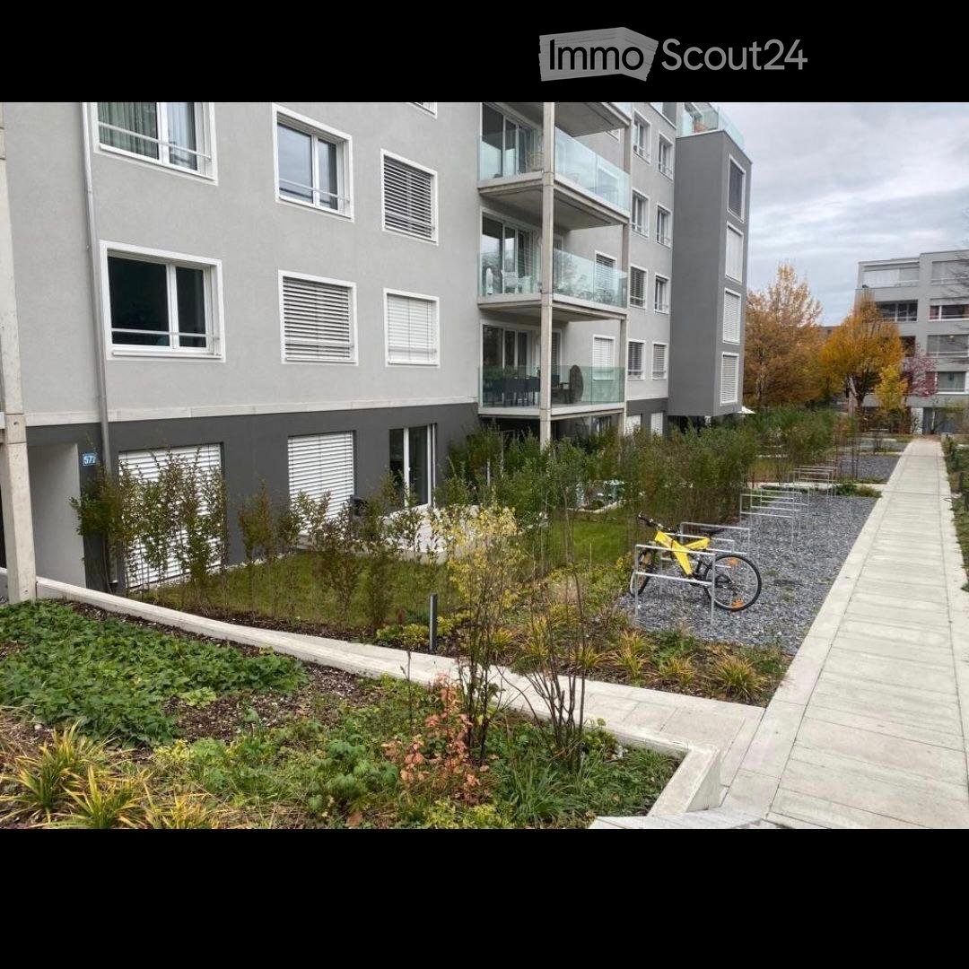 Modern multi-story apartment building with balconies, surrounded by landscaped greenery and a paved walkway.