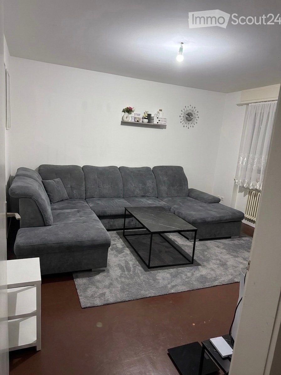 Modern living room with a grey sectional sofa, black coffee table, white shelf with decorative items, wall clock, side table, and curtained window.