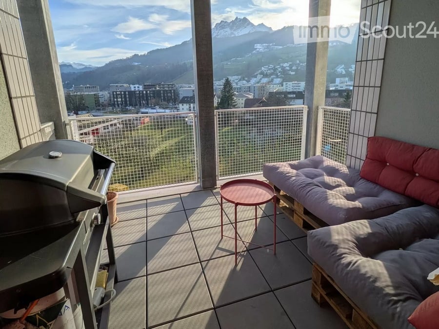 Balcony with a view of mountains and city, featuring a sofa, small round table, and grill.
