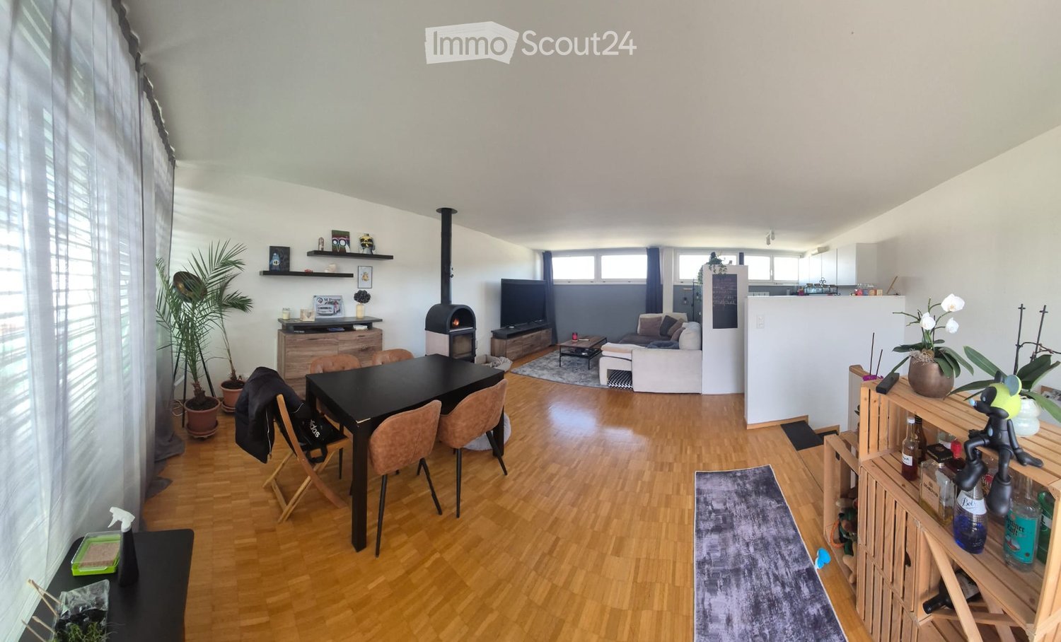 Open-plan living and dining area with wooden flooring, fireplace, sofa, coffee table, dining table with chairs, shelves, and plants.