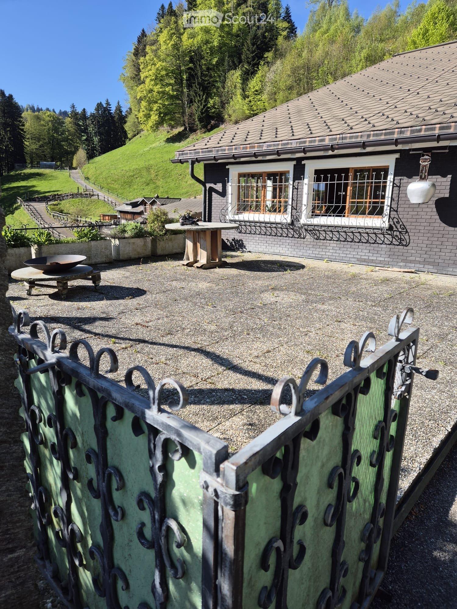 Outdoor patio area with gravel ground, metal fence, small table, bowl, and planters near a house with large windows and a sloped roof.