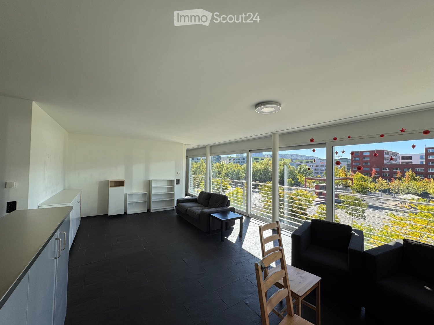 Modern living room with dark wooden flooring, white walls, large windows, and a balcony view. Features a sofa, coffee table, wooden chair, and built-in cabinets.