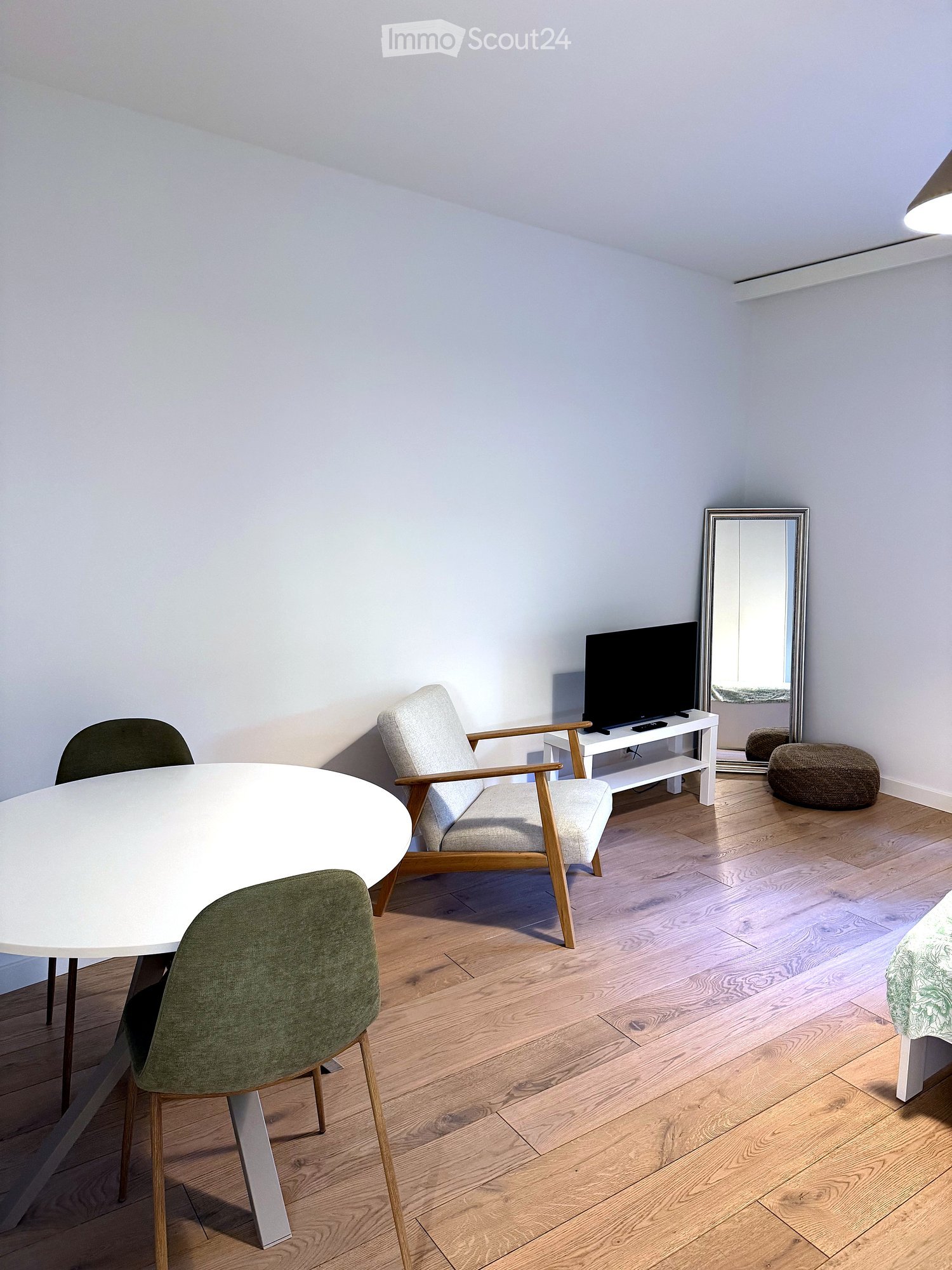 A modern, well-lit room with light wooden flooring, a white dining table with green chairs, a grey armchair, a television on a white stand, a round stool, and a large mirror on the wall.