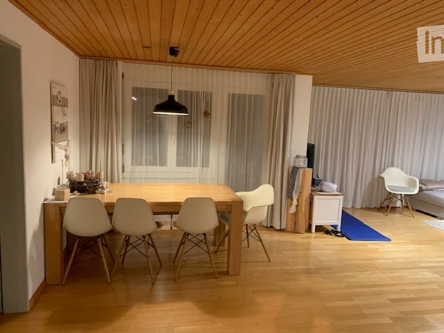 Dining room with wooden floor, dining table, four chairs, pendant lamp, and a TV screen in the background.