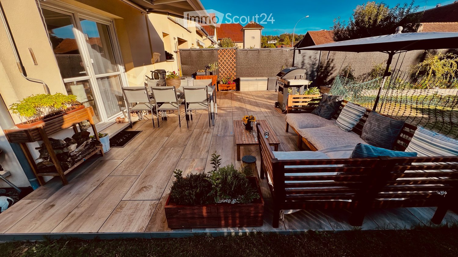 A wooden deck patio with a sofa, coffee table, chairs, umbrella, grill, and potted plants.