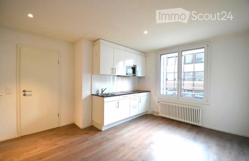 Empty kitchen with white cabinets, a sink, and a microwave. The space features wooden flooring, large windows, and a radiator under the windows.