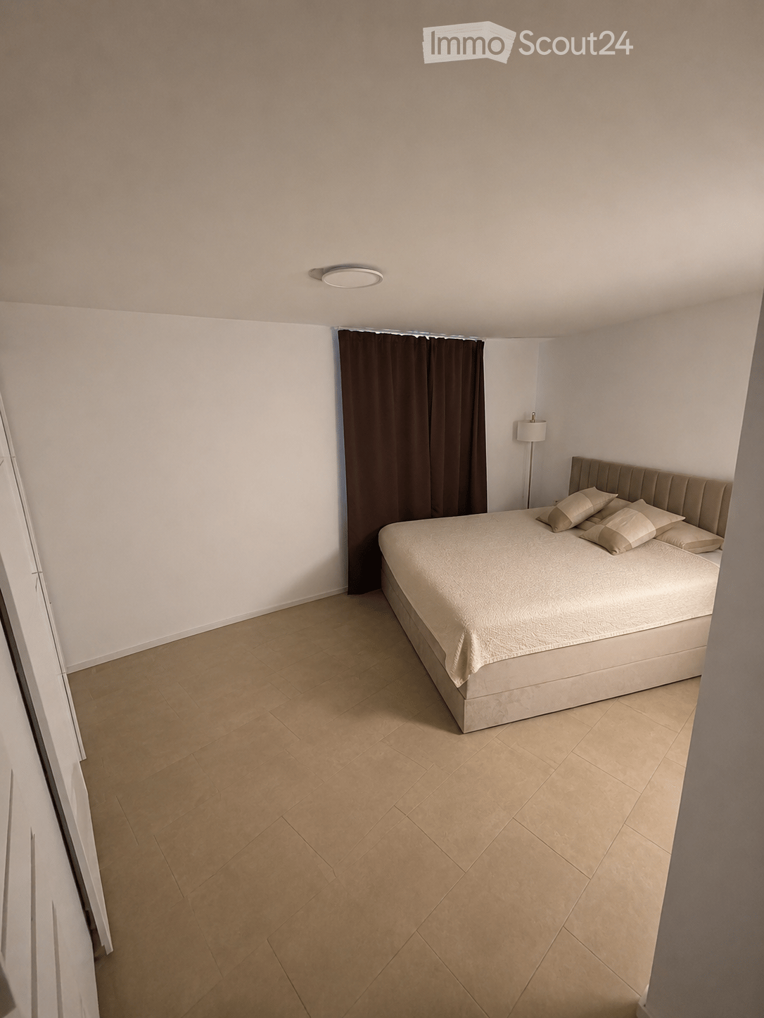 Modern bedroom with a large bed, beige pillows, a dark curtain, and a floor lamp near the window.