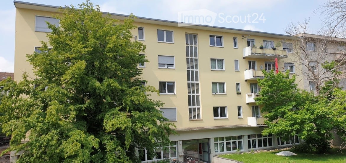 Four-story residential building with beige facade, multiple windows, and balconies on the upper floors.