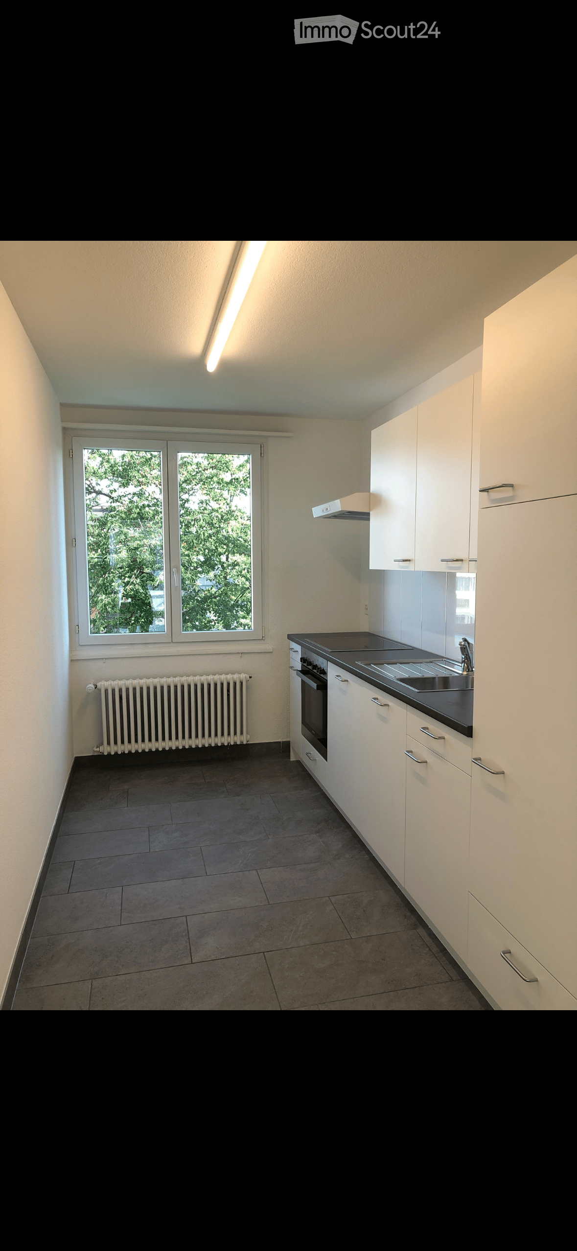 Empty kitchen with white cabinets, dark countertops, a sink, and a window overlooking greenery. The floor is tiled, and there is a radiator below the window.