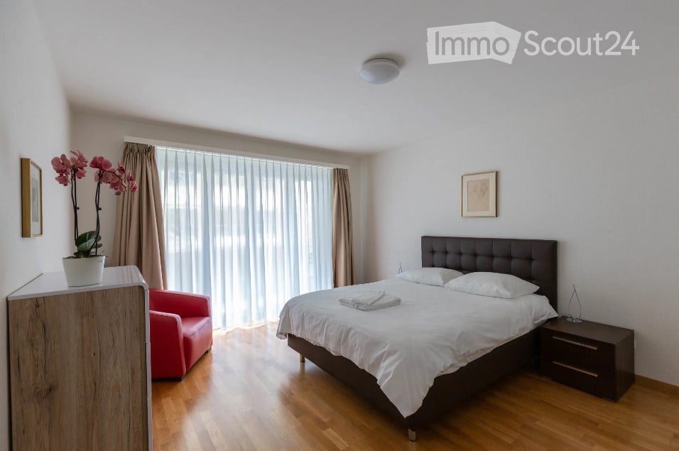 Modern bedroom with a large bed, nightstand, red armchair, flower pot, and large window with curtains.