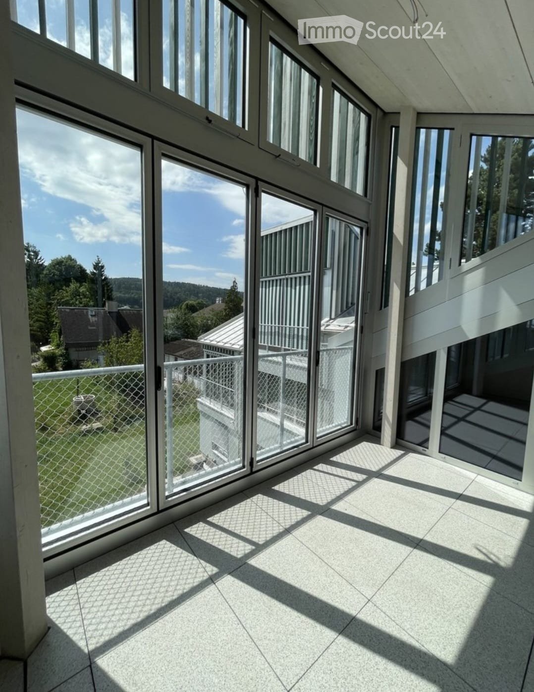 Bright, modern balcony with large glass windows and doors, white tiled floor, metal railing, and a view of a green lawn and trees.