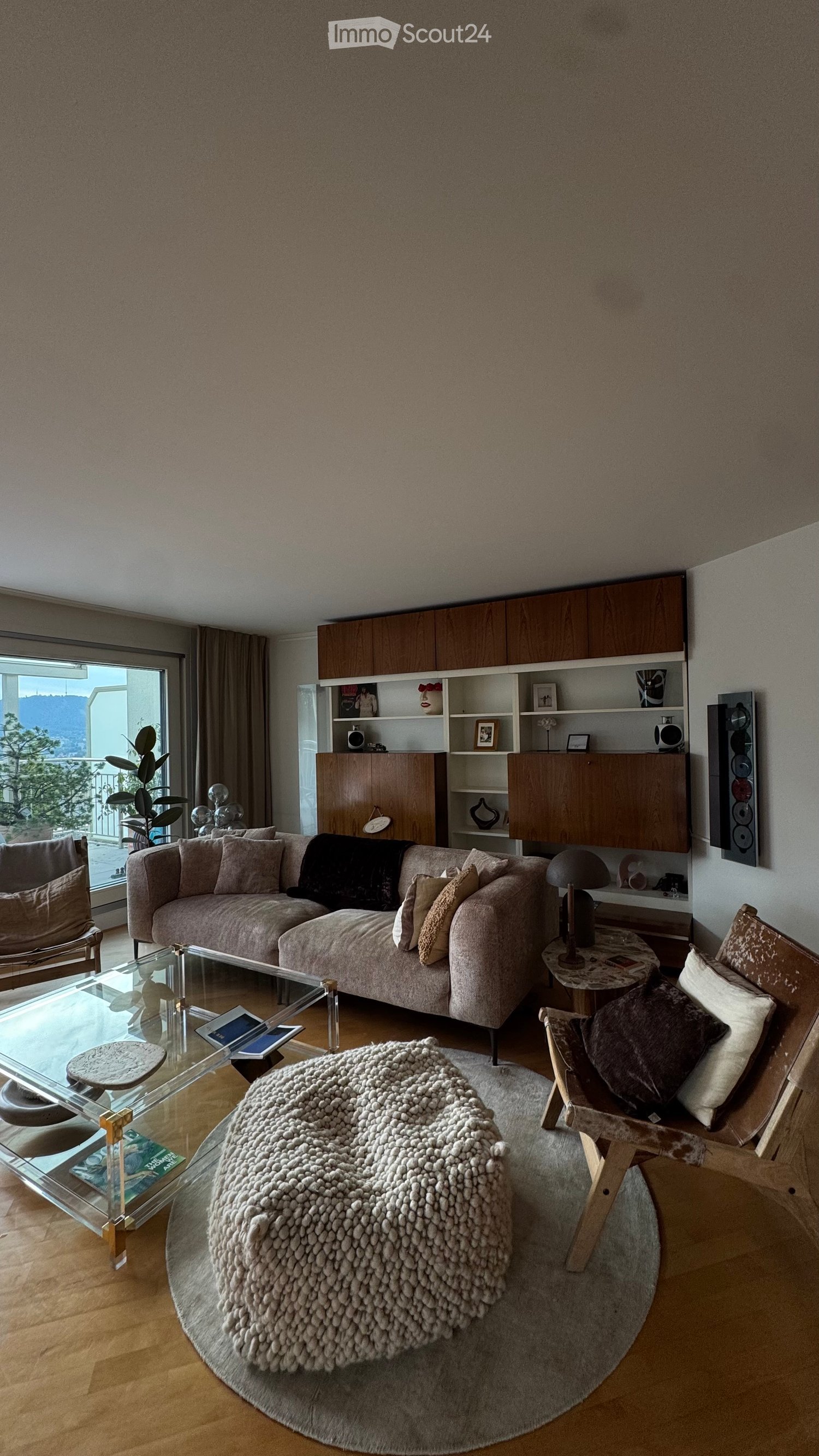 Modern living room with a large window offering a scenic view, featuring a beige sectional sofa, glass coffee table, round ottoman, wooden chairs, and a floor lamp.