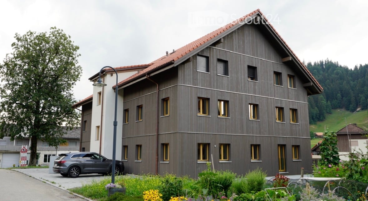 Modern two-story building with a sloped roof and wooden facade, surrounded by a garden with flowers and a parked car.