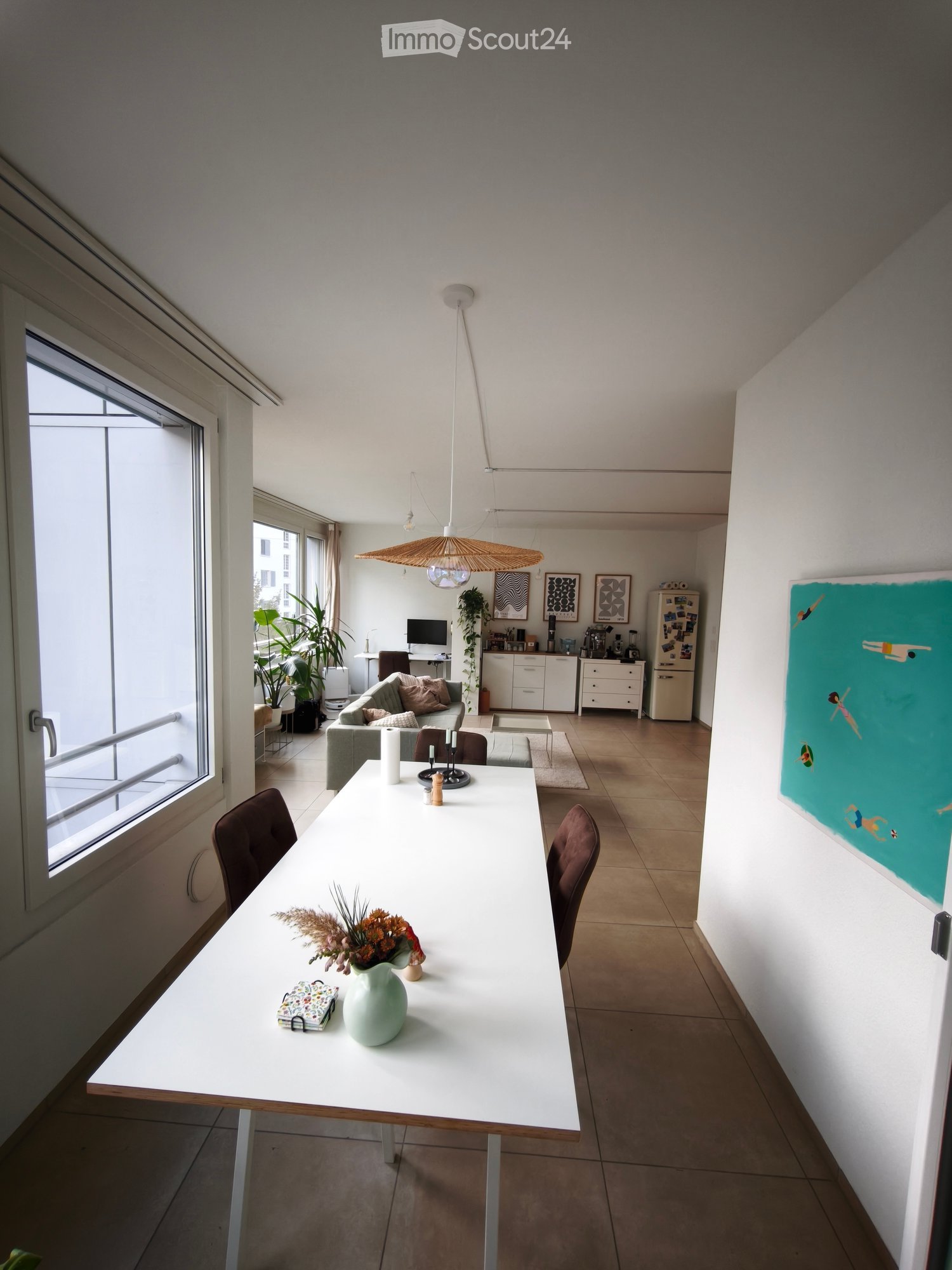 Open-plan living and dining area with a white dining table, chairs, a grey sofa, a refrigerator, and a large window with a view.