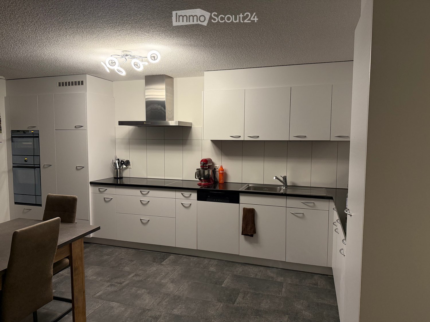 Modern kitchen with white cabinets, black countertops, and stainless steel appliances, including an oven, dishwasher, and mixer. A small dining area with a table and chairs is visible.