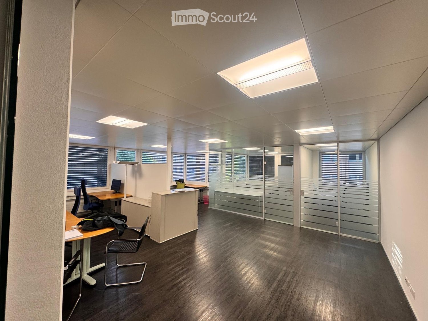 Modern office space with dark wooden flooring, glass partitions, desks, chairs, and computer screens.