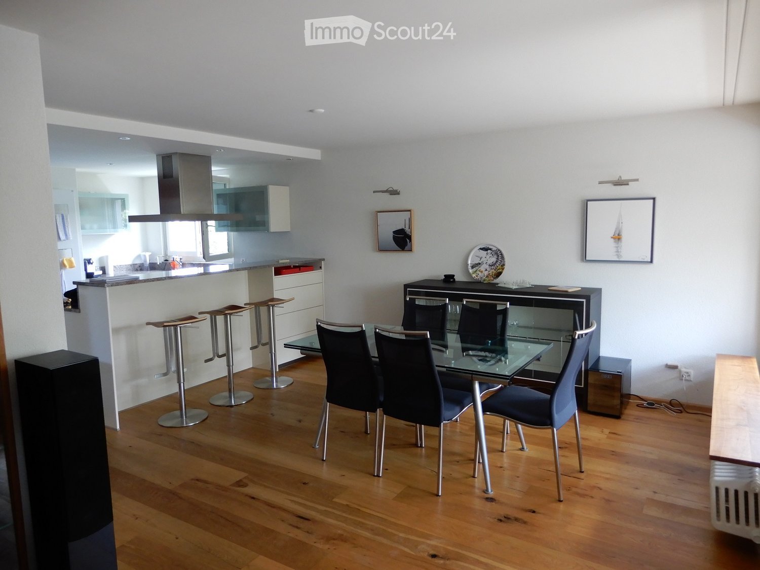 Modern dining area with a glass dining table, six chairs, four bar stools, a kitchen island, and framed artwork on the wall.