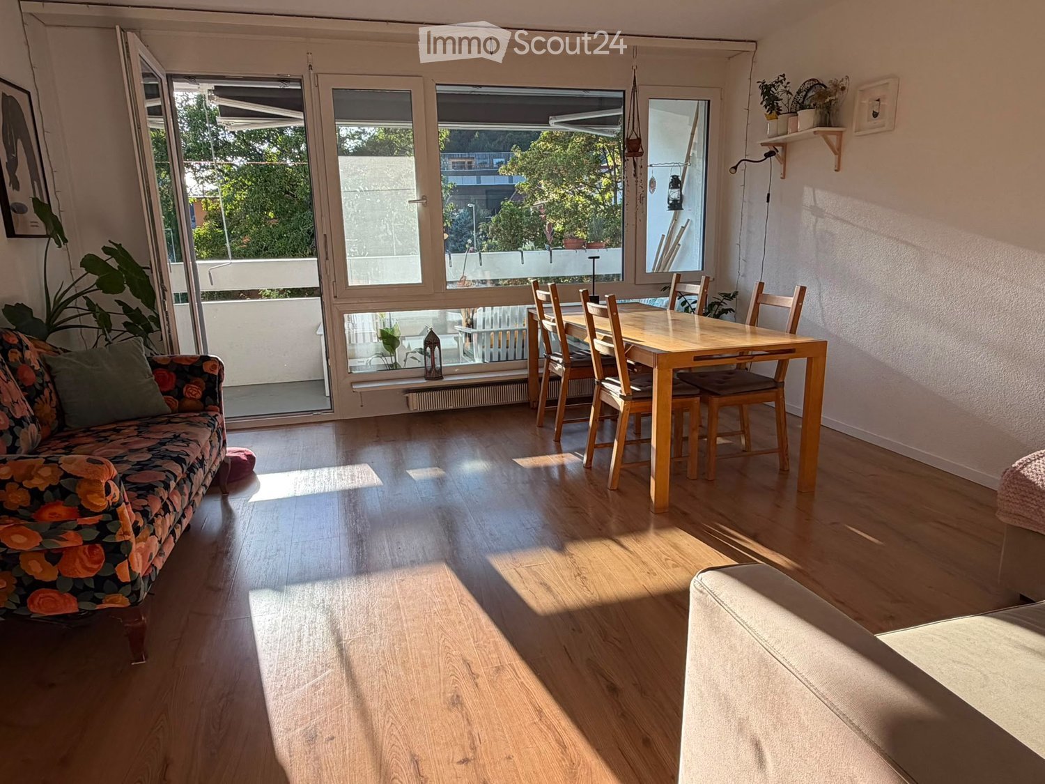 Bright living room with wooden flooring, large windows, a floral-patterned sofa, a wooden dining table with chairs, and potted plants.