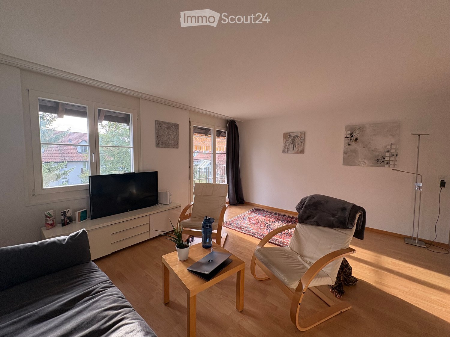 Modern living room with light pink walls, wooden flooring, large windows, a sofa, coffee table, chairs, a laptop, a television, and a floor lamp.
