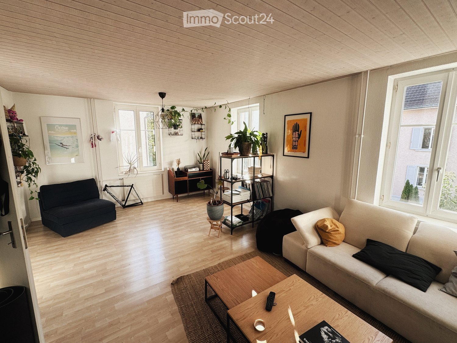 Modern living room with light wooden flooring, white walls, and large windows. Features a cream-colored sofa with pillows, a dark-colored sofa, a wooden coffee table, a shelf with books and plants, and several potted plants around the room.