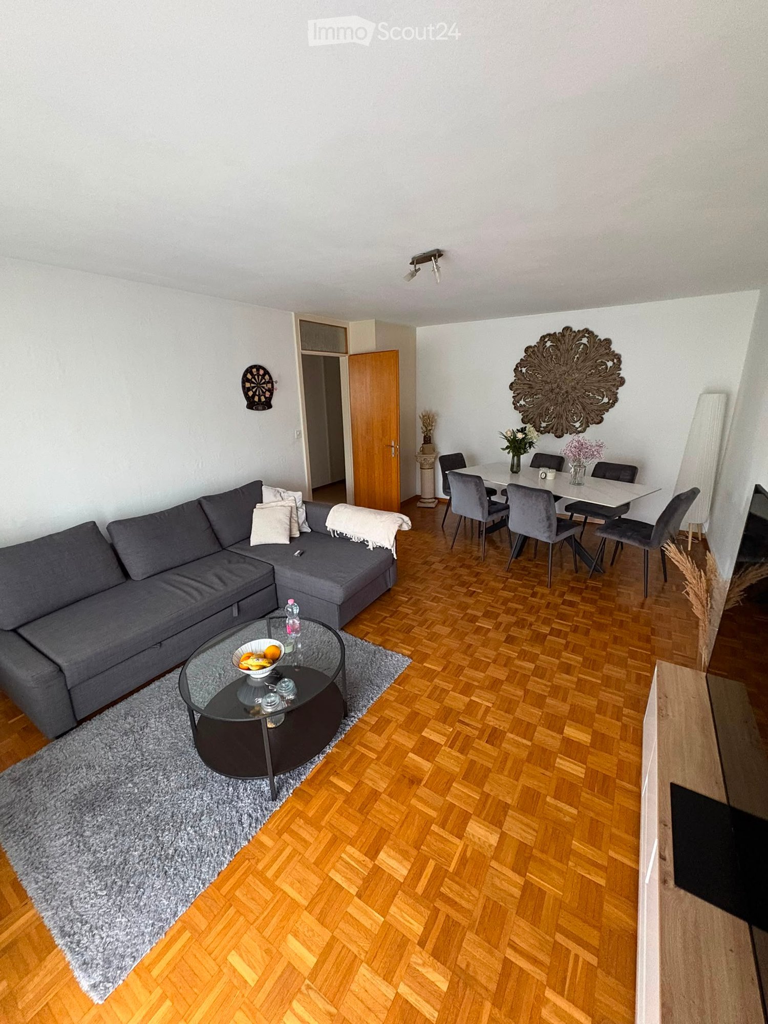 Living room with grey sectional sofa, glass coffee table, dining area with white table and grey chairs, decorative mirror, and dartboard on wall.