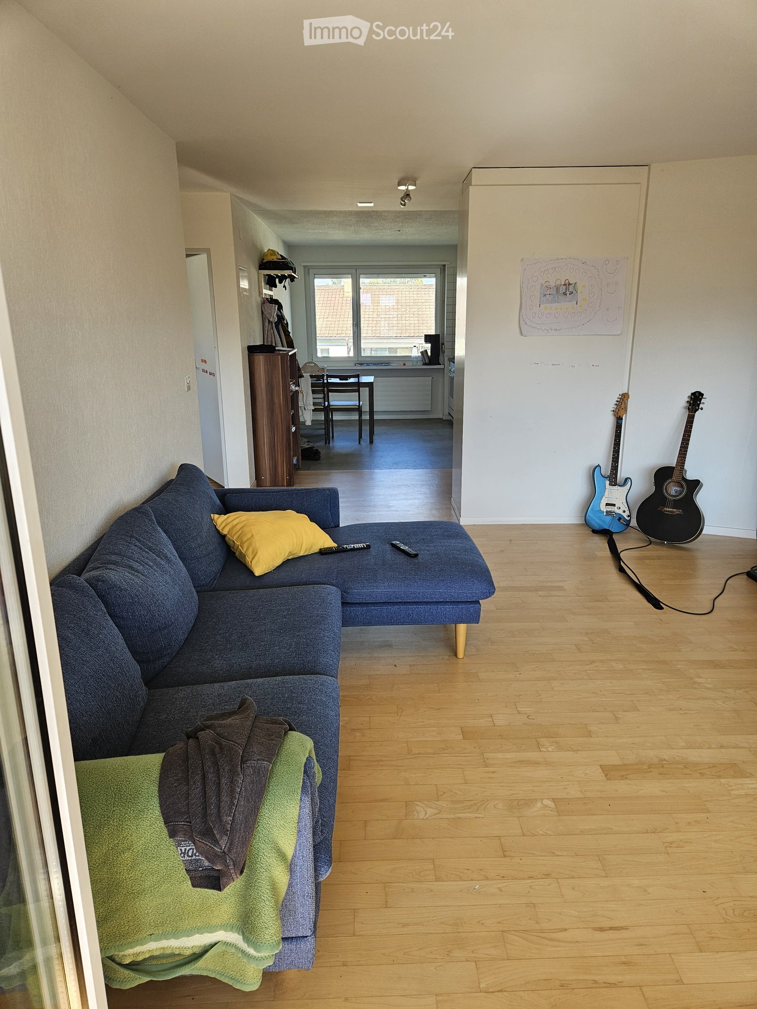 A compact, furnished room with a sofa, guitar on the wall, a small table, and a window providing natural light.