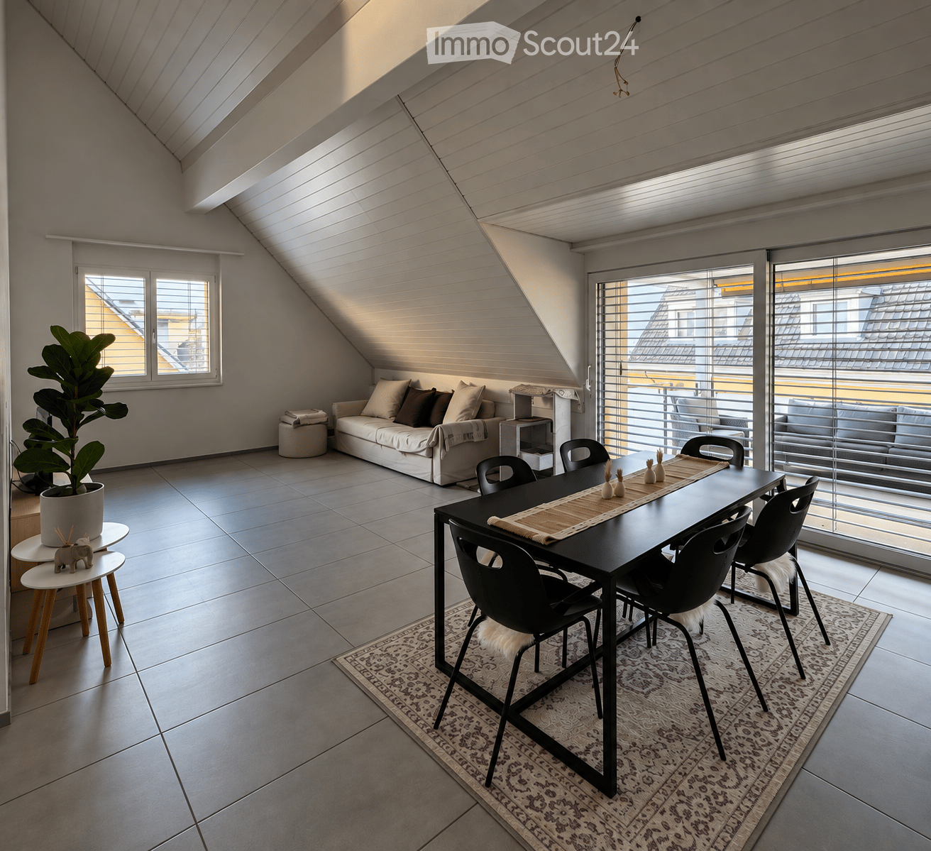 Modern attic room with slanted ceilings, large windows, and a balcony. Features a white sofa, black dining table with chairs, small wooden stools, a large potted plant, and a patterned rug.