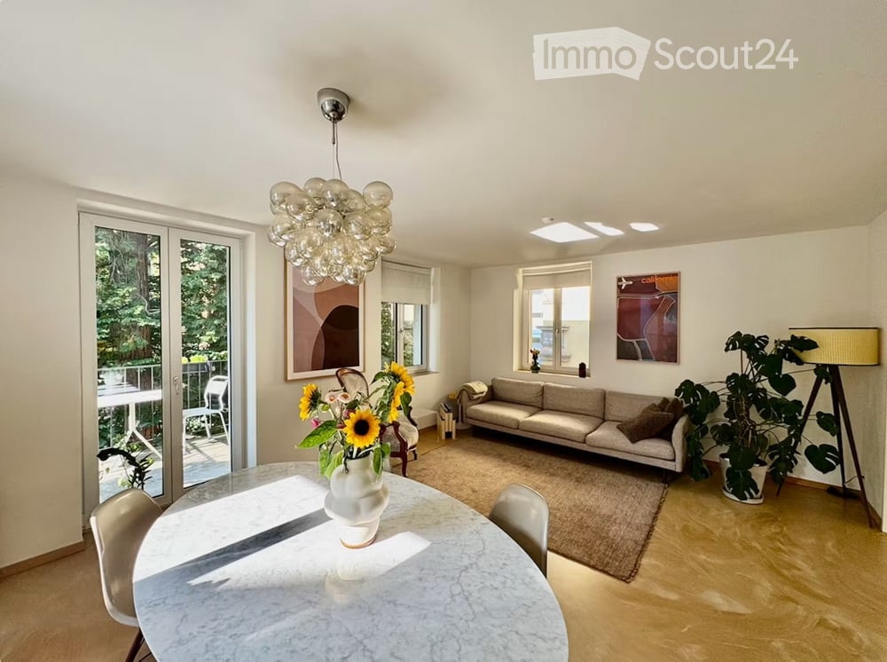 Bright living and dining area with a modern sofa, dining table with chairs, a large chandelier, and a view of a balcony.