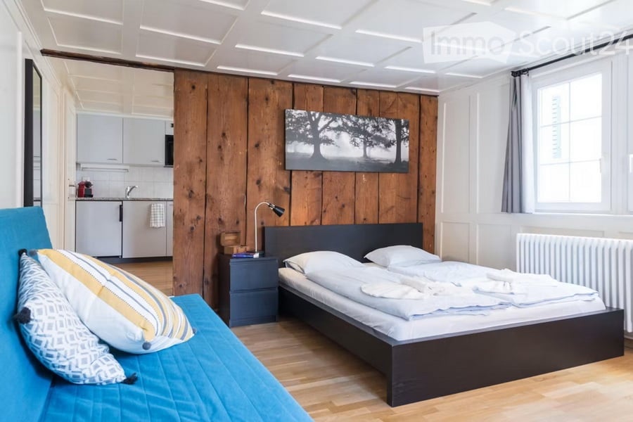 A modern, compact bedroom with a double bed, nightstand, sofa, and a small kitchen area with cabinets and a sink.