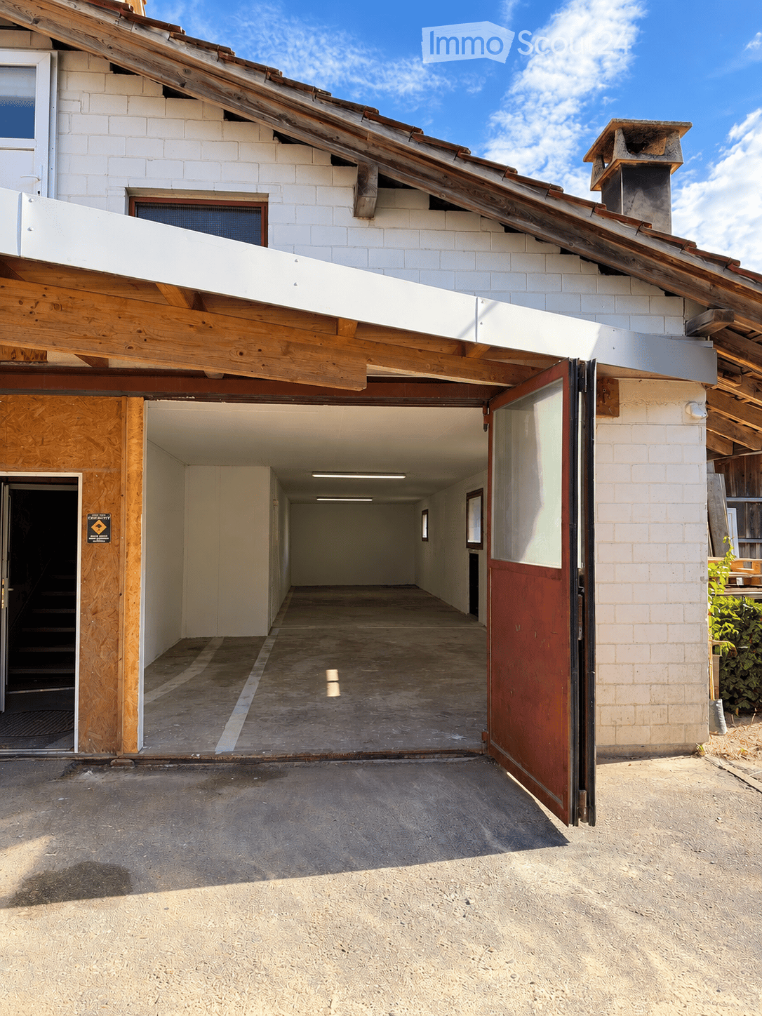 A modern building with a large garage entrance, white brick walls, wooden beams, and a slanted wooden roof. The garage has a concrete floor and is partially open, revealing a hallway inside.