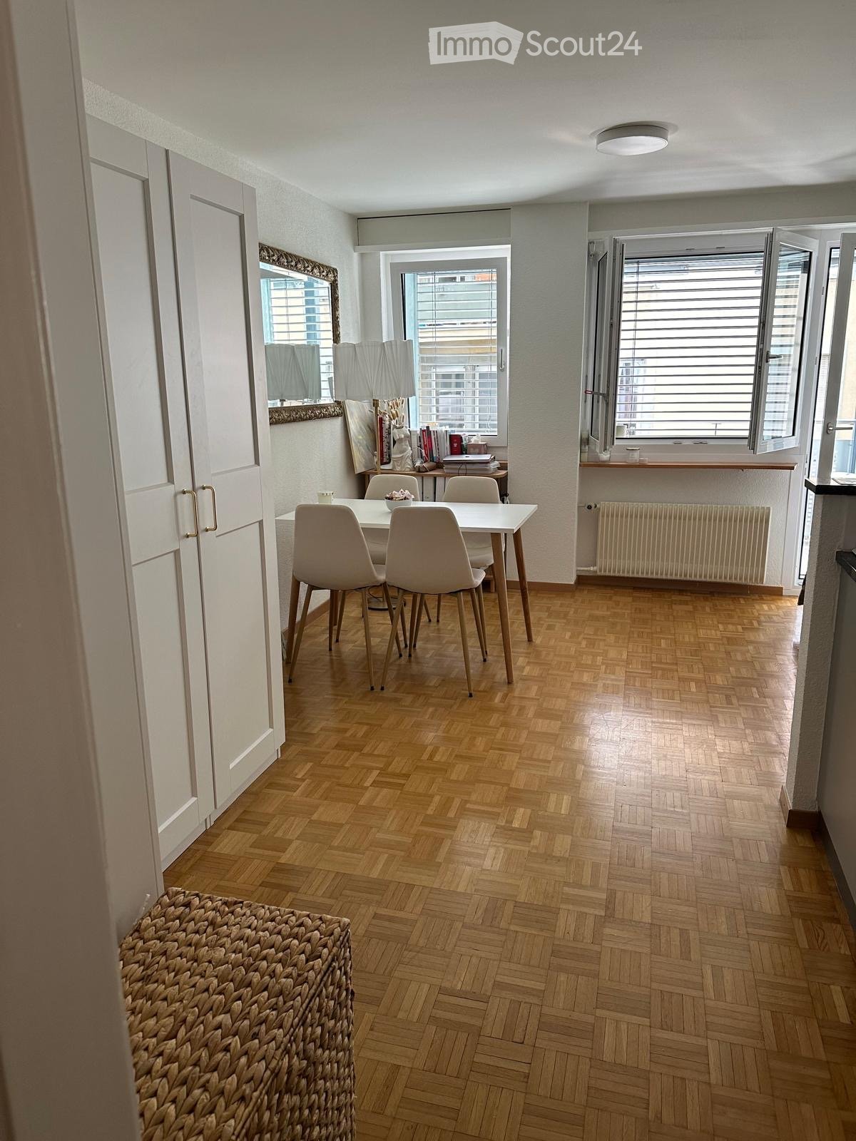 Dining area with wooden herringbone flooring, white table and chairs, large windows with blinds, radiator, and a woven basket on the floor.