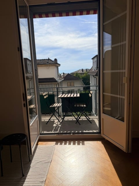 Balcony with a table and chairs, overlooking a cityscape with rooftops and distant hills under a clear blue sky.