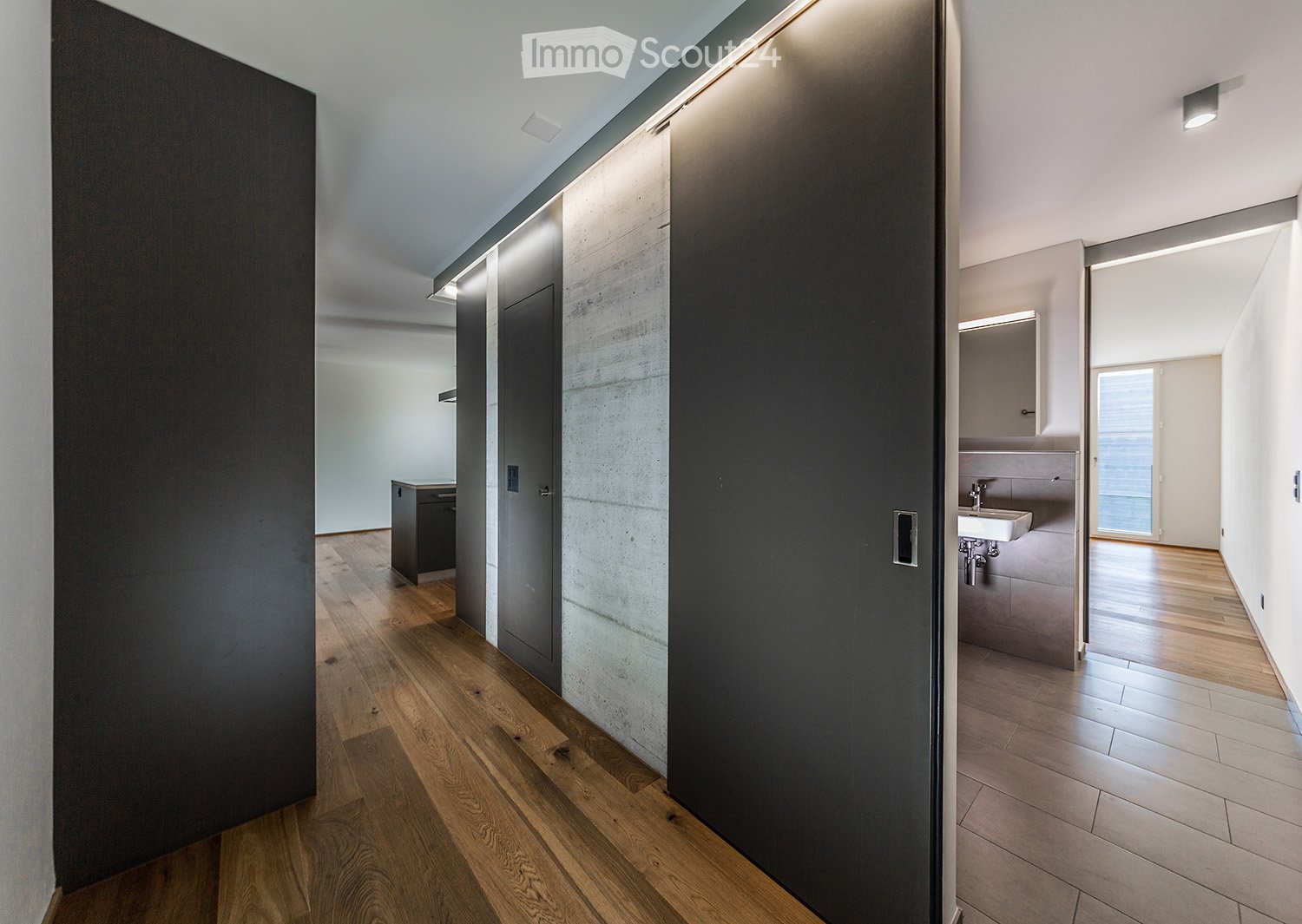 Modern hallway with wooden flooring, sleek black and grey walls, recessed lighting, and a visible bathroom area with a sink and mirror.