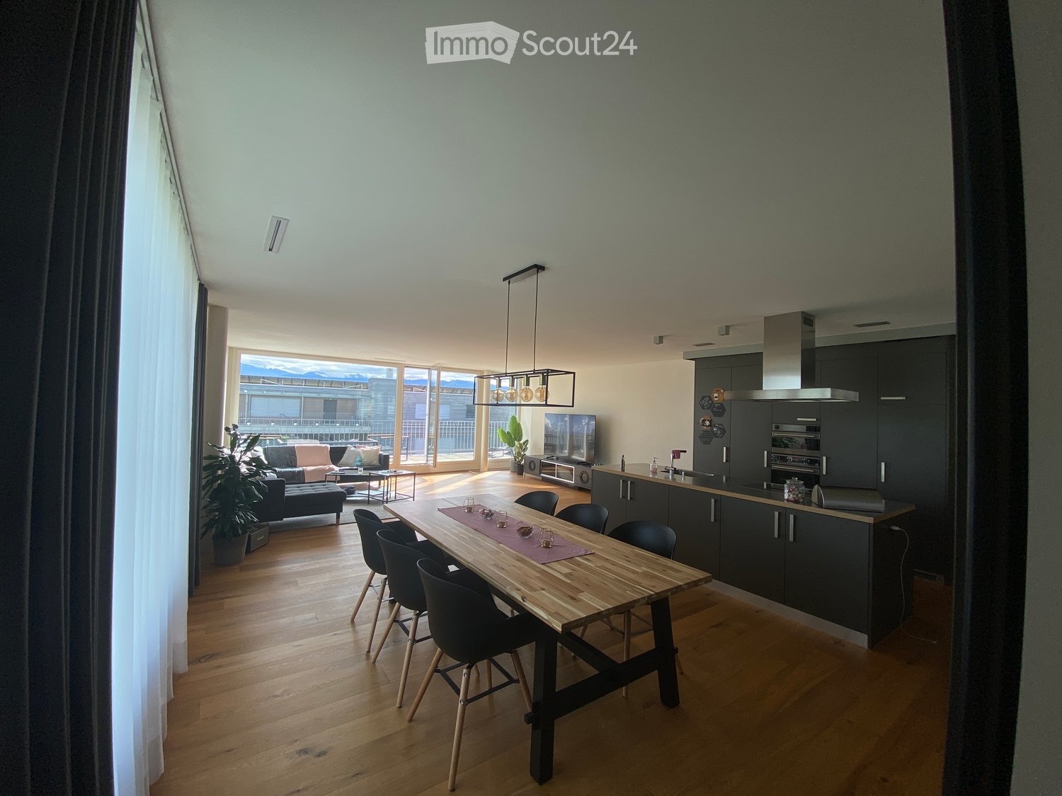 Open-plan living and dining area with modern furniture, including a sofa, coffee table, dining table with chairs, and a kitchen area with cabinets, appliances, and a range hood. Large windows offer a scenic view.