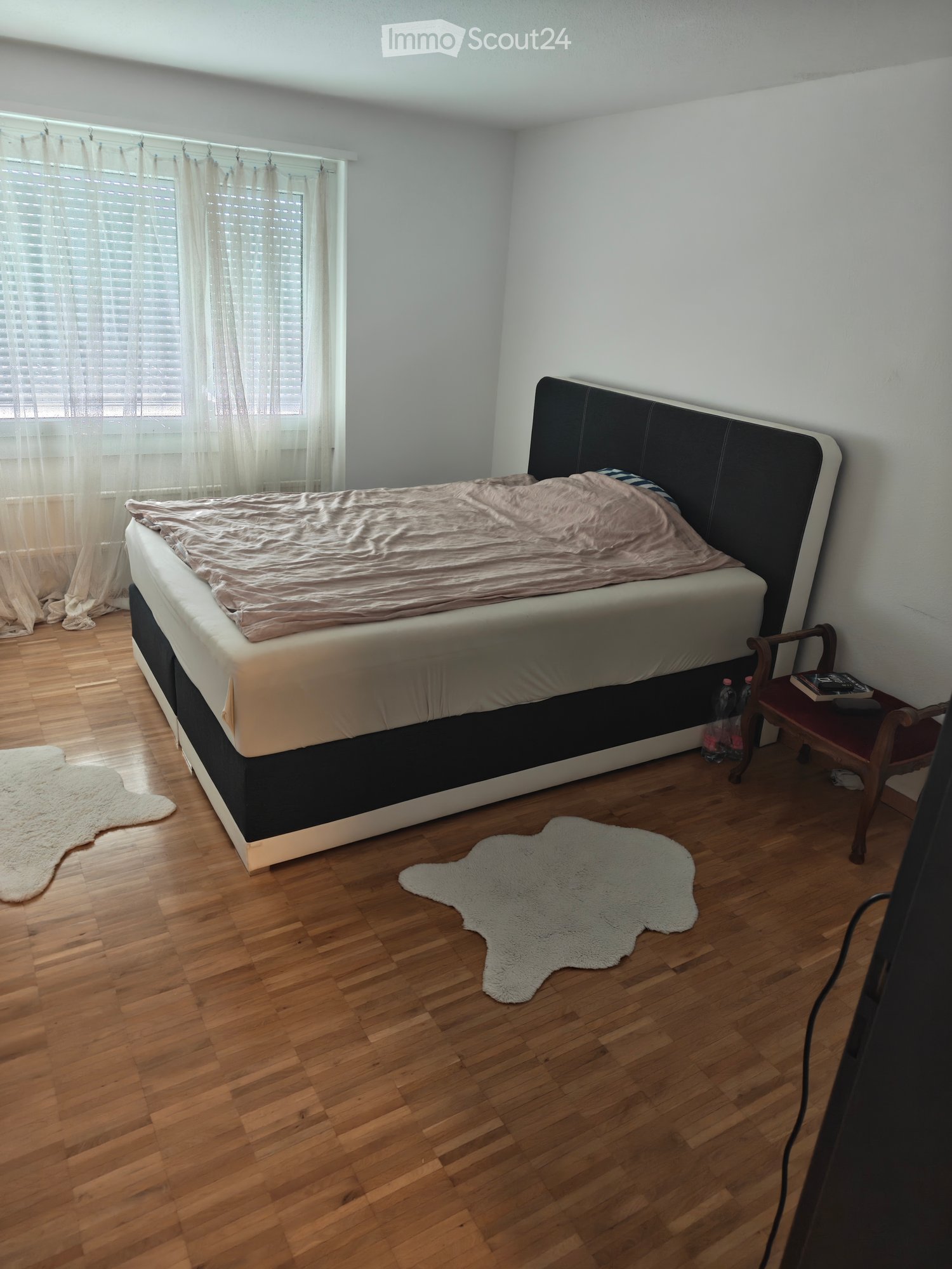 A well-lit bedroom with a large bed, a wooden chair, a small table, and two white rugs on the floor.