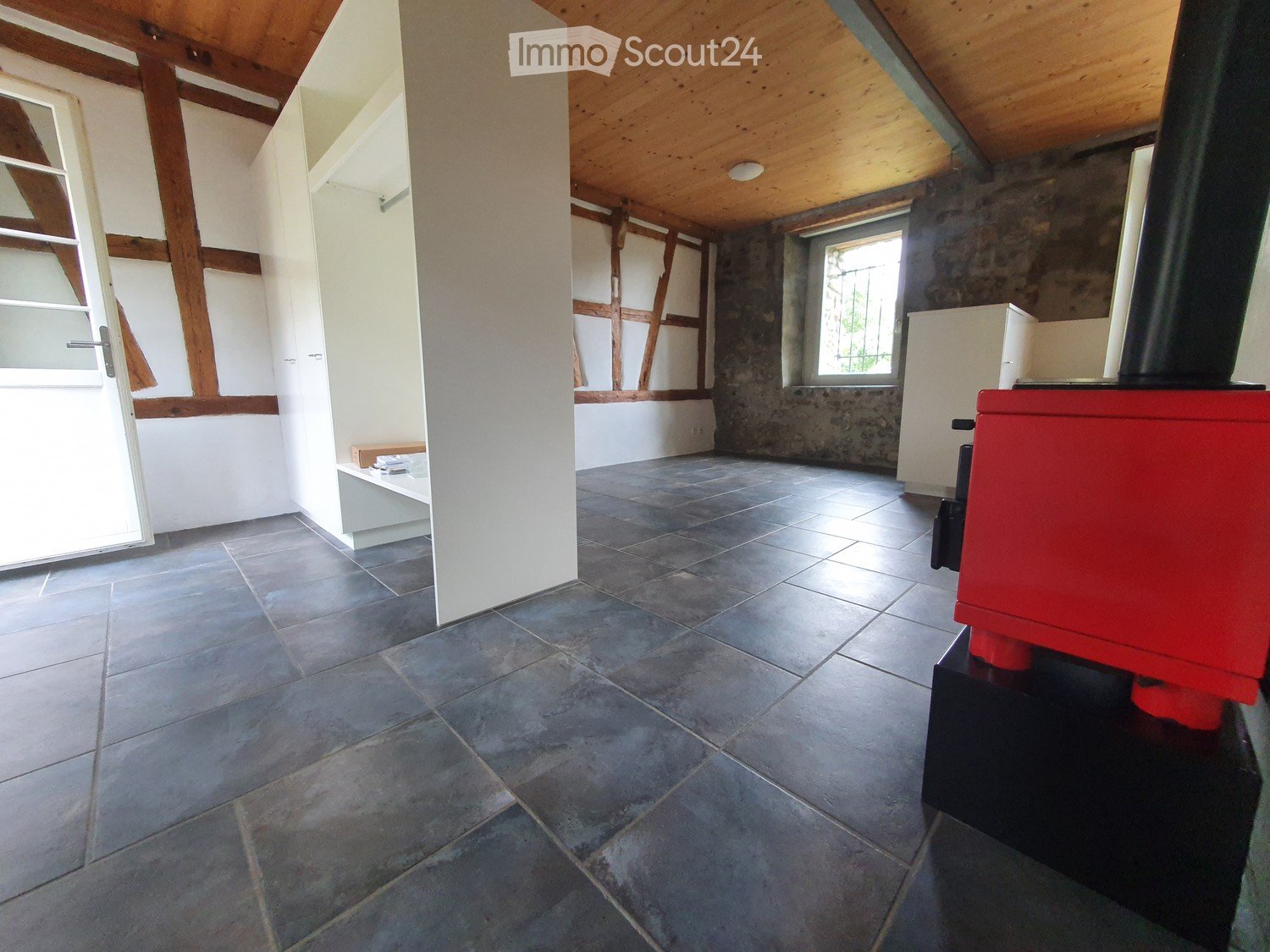 Empty room with exposed wooden beams, tiled floor, and large windows allowing natural light. Features include a small bench and a red stove in the corner.