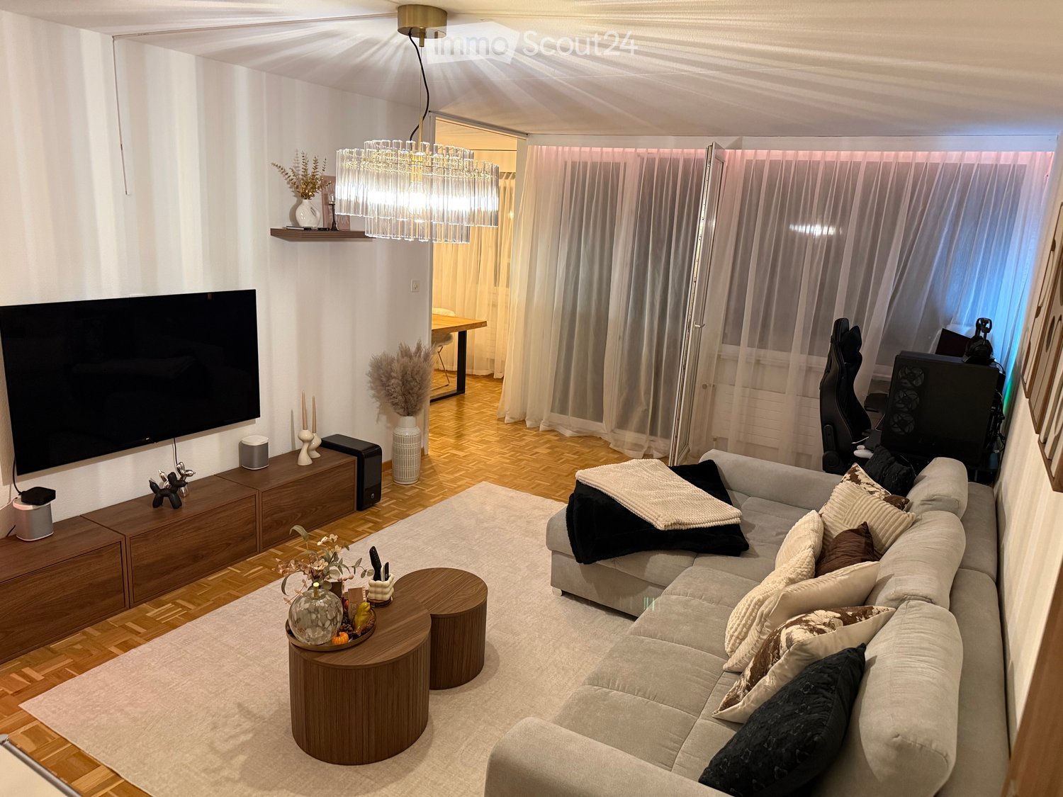 Modern living room with a grey sectional sofa, wooden coffee table, large flat-screen TV, and decorative vases. The space features wooden flooring, large windows with sheer curtains, and a chandelier hanging from the ceiling.