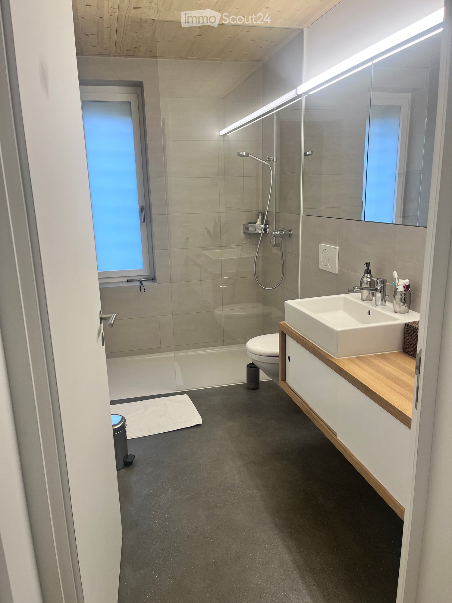 Modern bathroom with a white sink, wooden vanity, large mirror, shower area, and a toilet. Features include a sleek design with grey tiles and a dark grey floor.