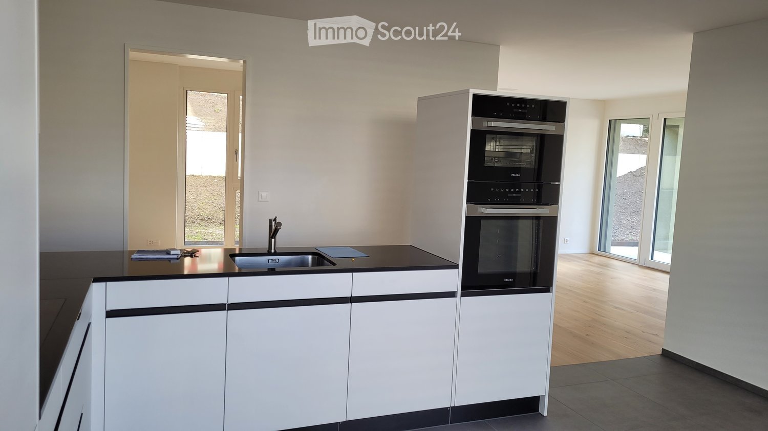 Modern kitchen with white cabinets, black countertops, a stainless steel sink, and a built-in oven. The space features large windows and a clean, minimalist design.