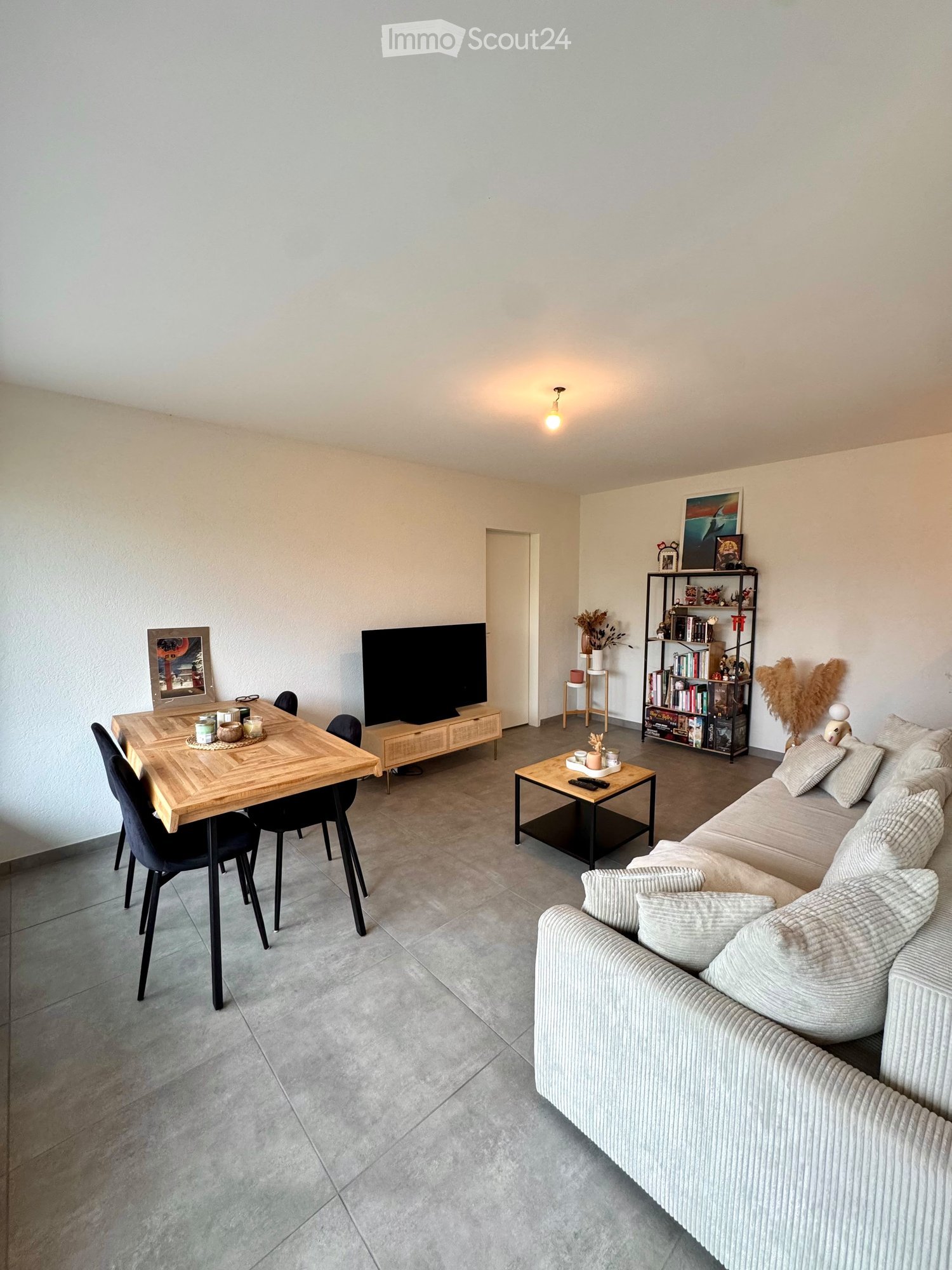 Modern living room with a grey sectional sofa, coffee table, dining table with chairs, bookshelf, and television. Neutral tones and contemporary decor.